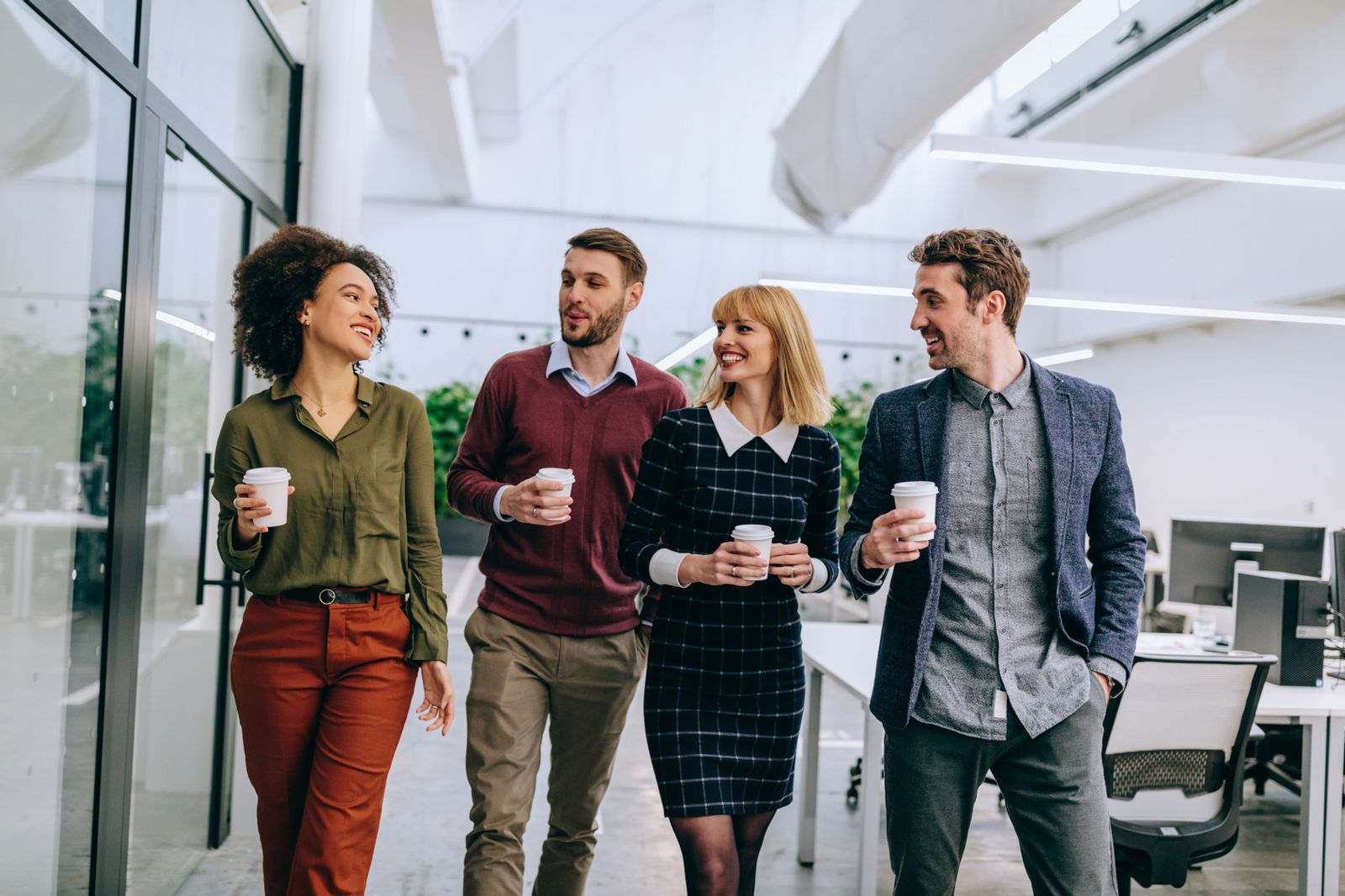 Four professionals walk together in a modern office, each holding coffee cups and smiling, dressed in smart casual attire.
